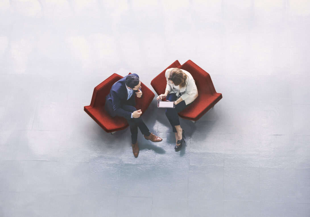 A high-angle view of a businessman and a businesswoman sitting in the office building lobby and using a tablet computer